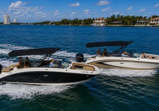 Two luxury motorboats cruising side by side on clear blue Miami waters near waterfront mansions.